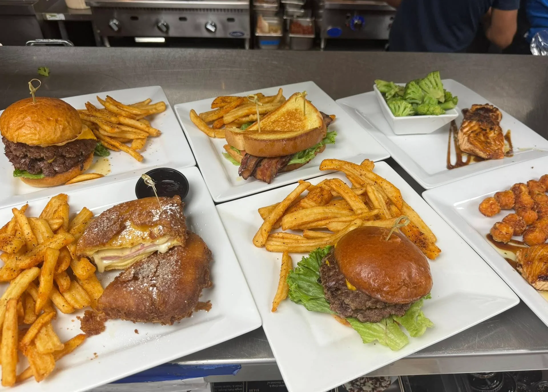 An assortment of plates including burgers with fries, grilled salmon with broccoli, tater tots, and a Monte Cristo sandwich, ready to be served.