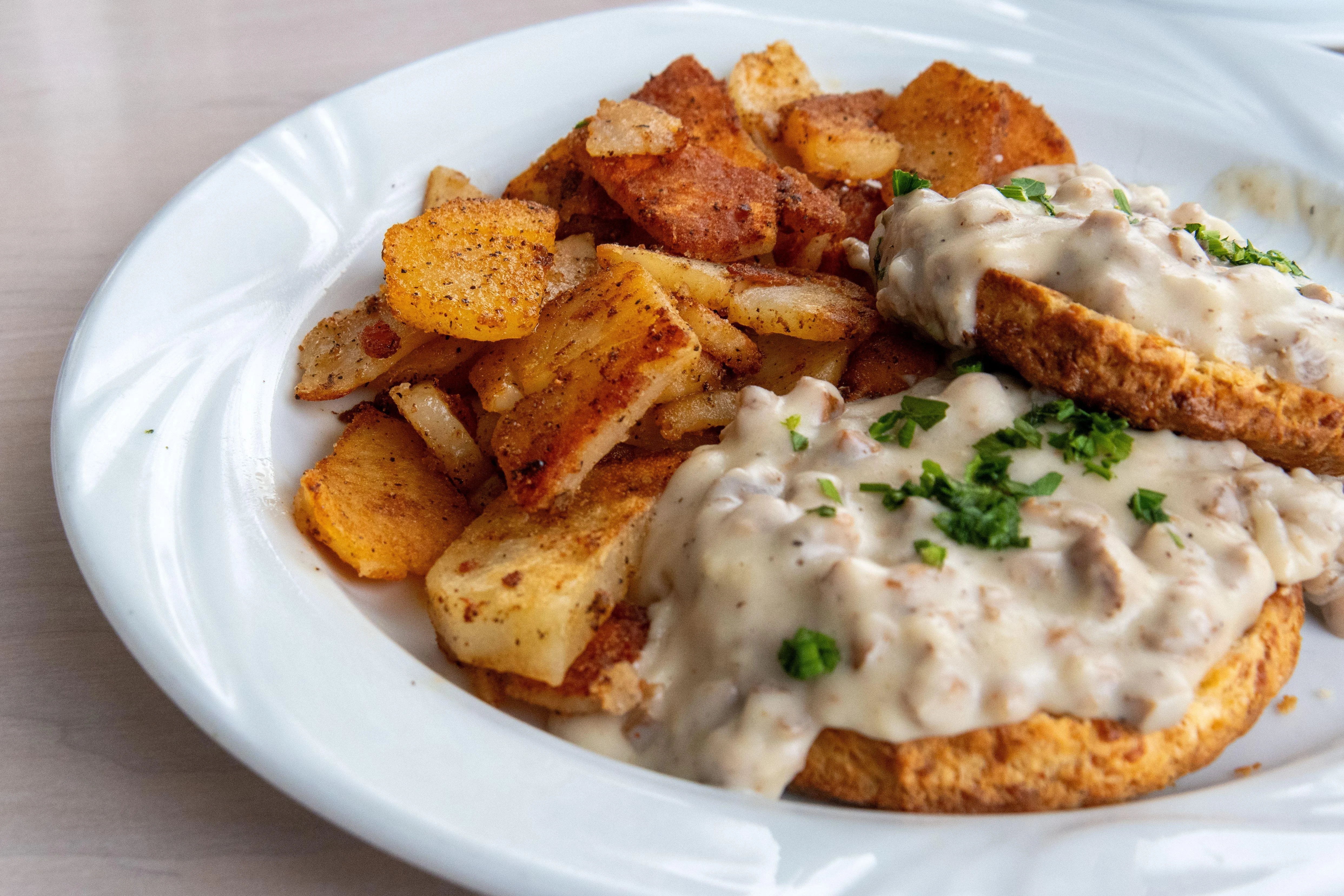 A hearty dish of biscuits smothered in sausage gravy, served alongside crispy golden brown potatoes. Arranged on a white plate.