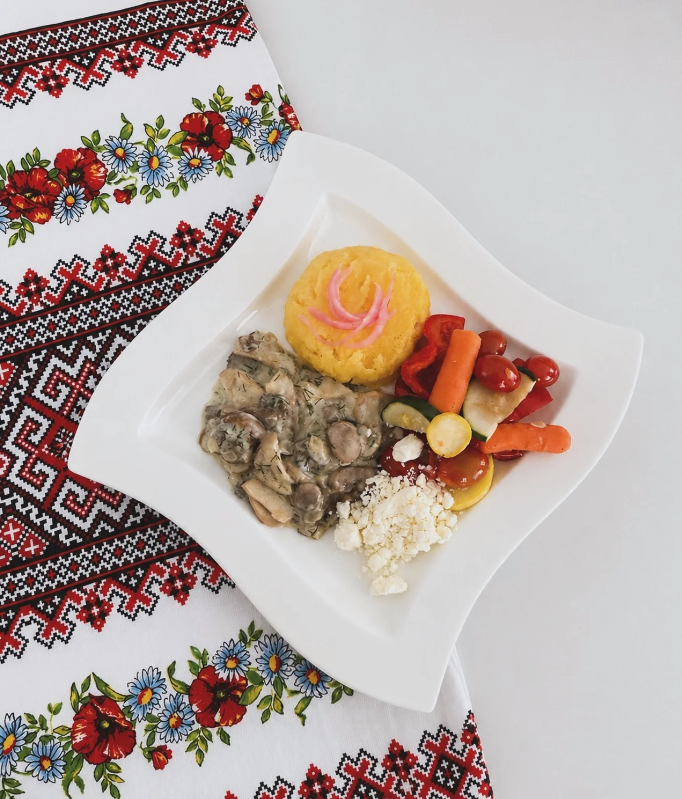 A plate featuring a serving of polenta, accompanied by a creamy mushroom dish, mixed vegetables, and fresh cheese, all arranged on a patterned tablecloth.