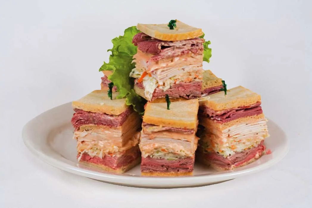A plate of stacked sandwiches made with layers of various meats and vegetables, garnished with a leaf of lettuce and served on a white plate.