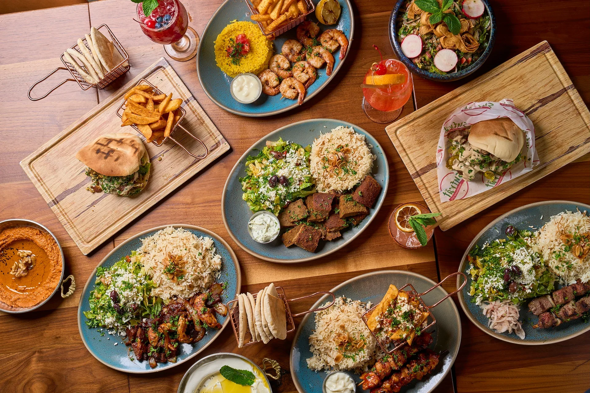 An inviting spread of Mediterranean dishes on a wooden table, featuring grilled meats, rice, pita bread, vibrant salads, and colorful drinks.