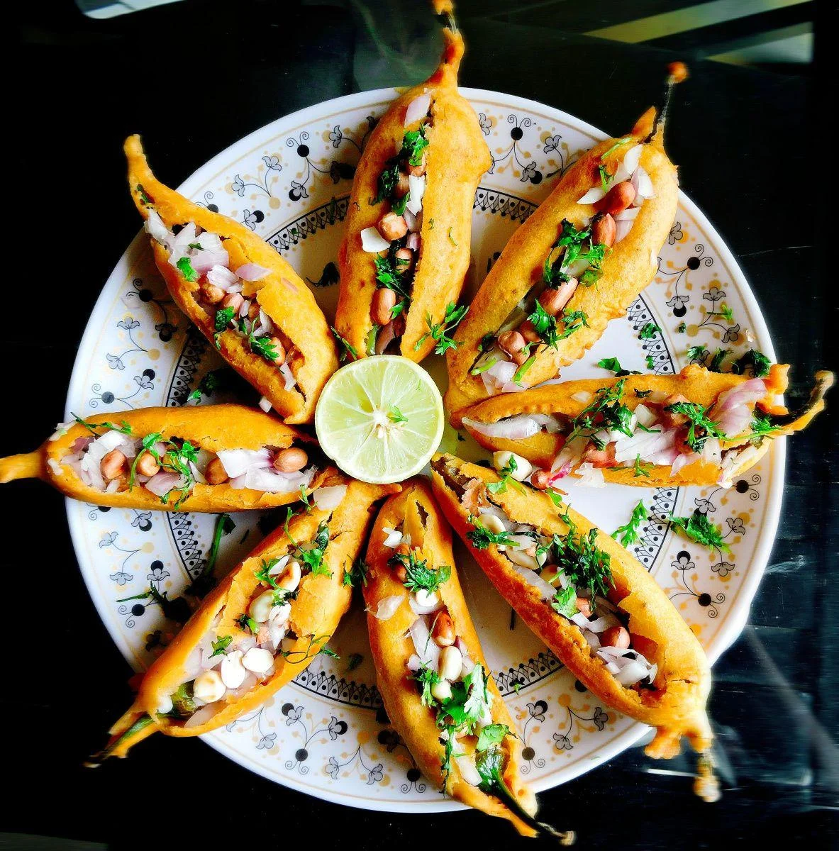 A platter of stuffed fried snacks arranged in a circular pattern, topped with chopped onions, peanuts, and a lime in the center.