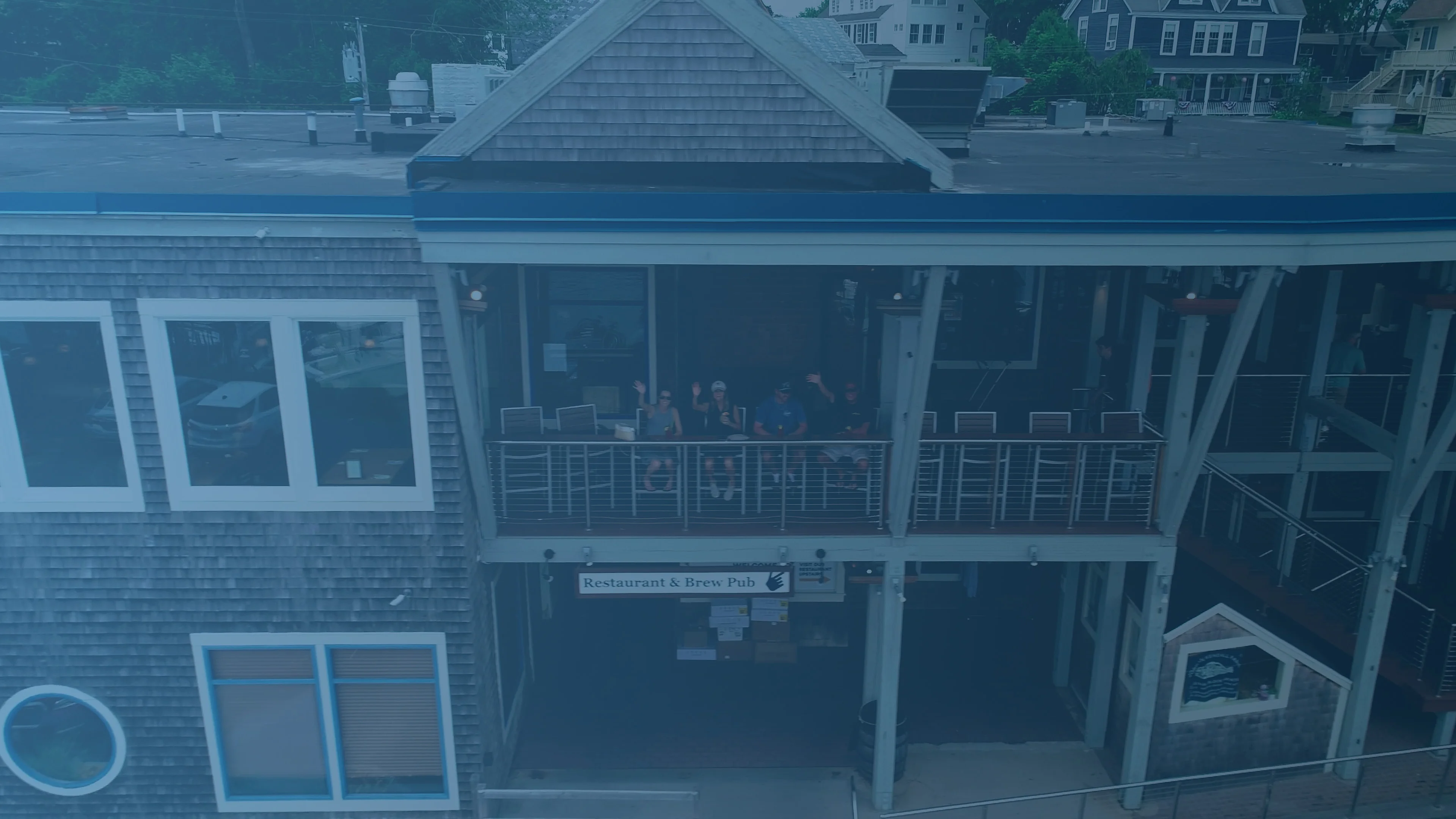 An aerial view of a restaurant with a deck, where people are seated and waving. The structure features large windows and a sign indicating it's a brew pub.