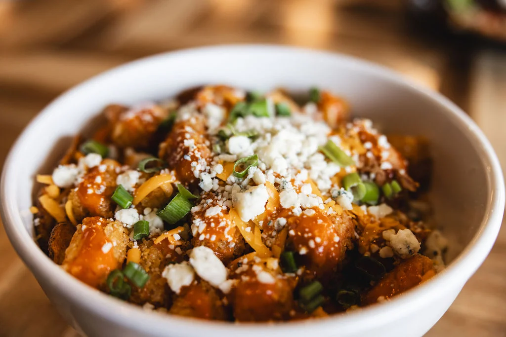 A bowl of loaded tater tots topped with cheese, crumbled feta, and green onions.