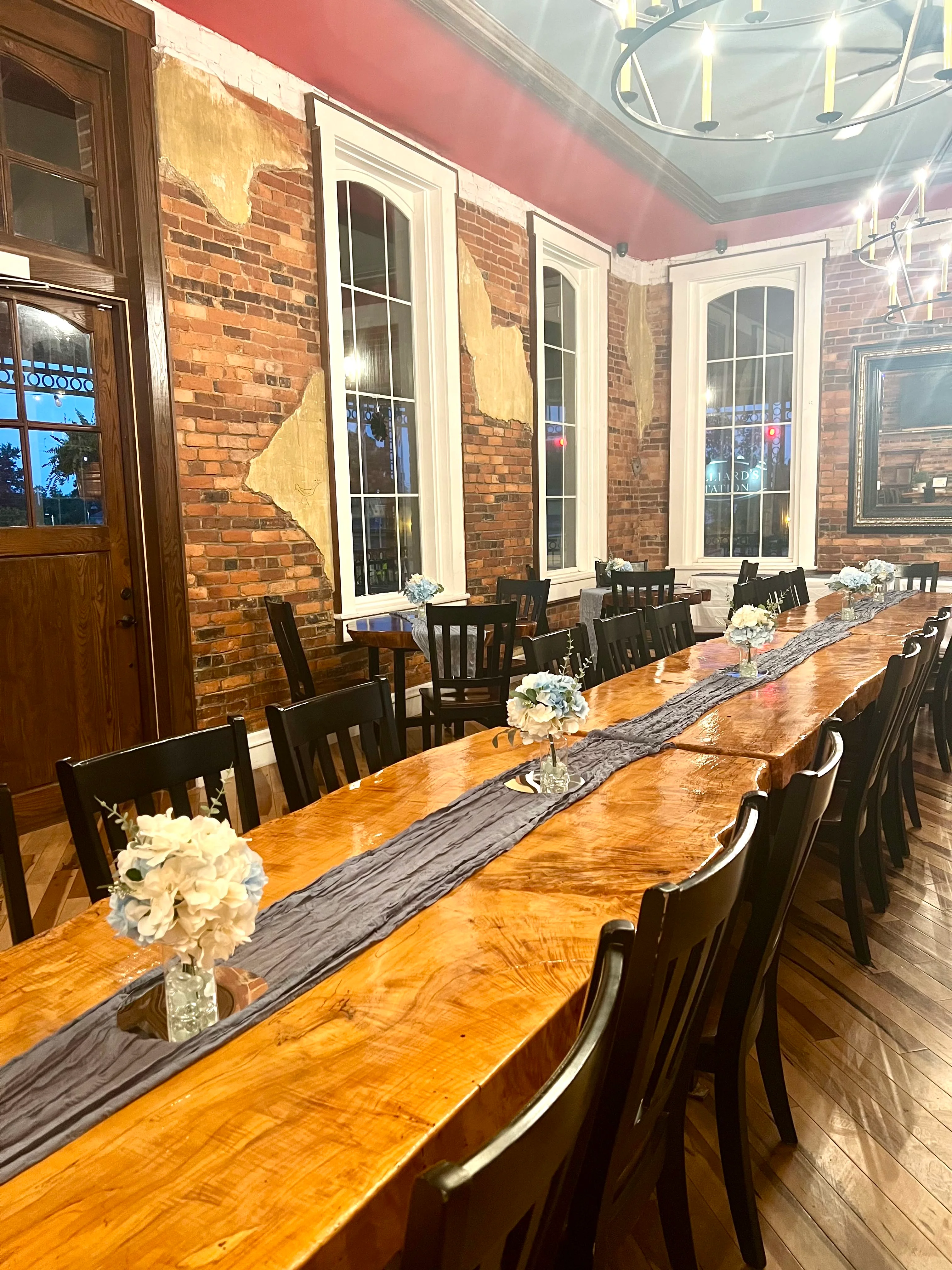 A long wooden dining table adorned with floral arrangements and a decorative runner, set in a stylish indoor venue with brick walls.