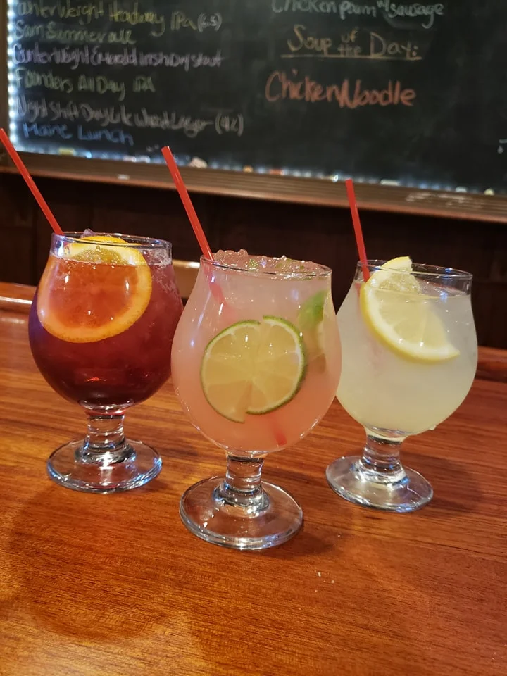 Three colorful cocktails served in decorative glasses, garnished with citrus slices, on a wooden bar top.