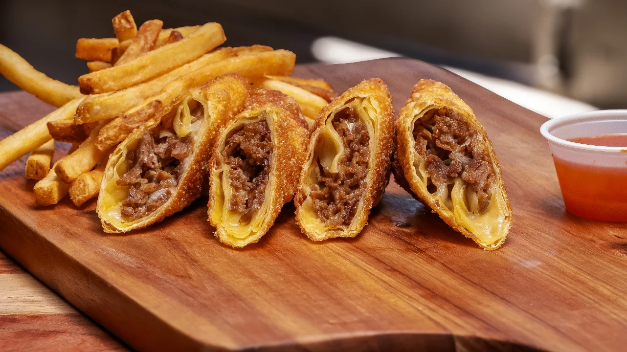 Three crispy beef-filled pastries served alongside French fries and a small cup of dipping sauce on a wooden board.