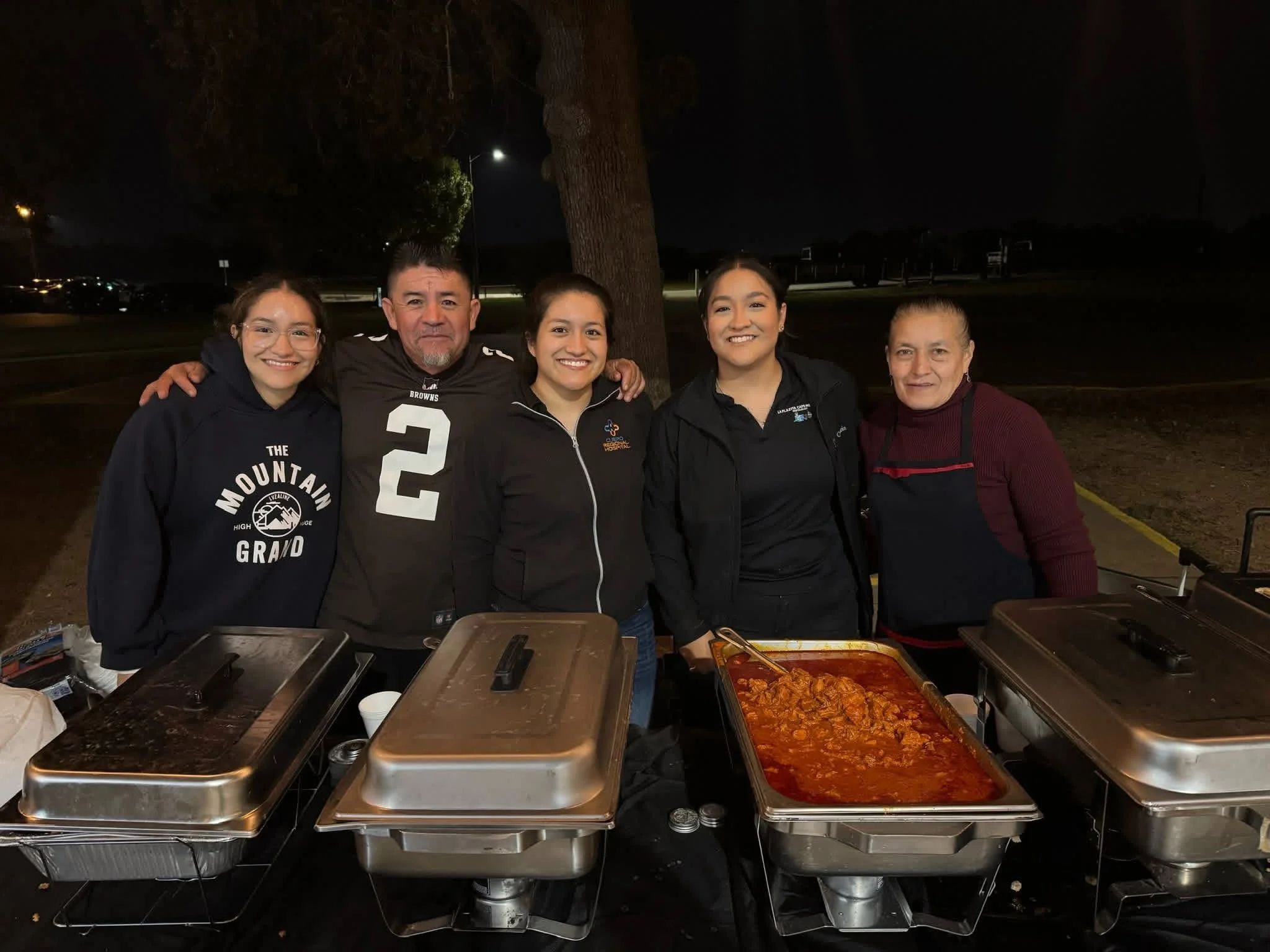 Group of people smiling behind a food serving table at night.