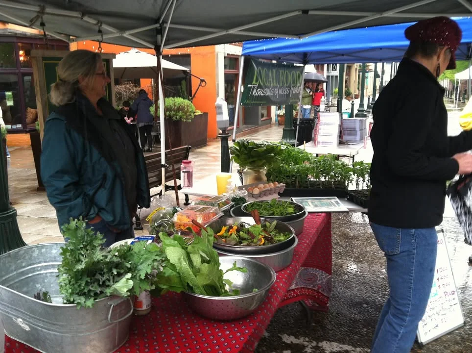 A vibrant market scene with fresh greens and vegetables displayed on a table under a tent, with vendors engaging with customers.