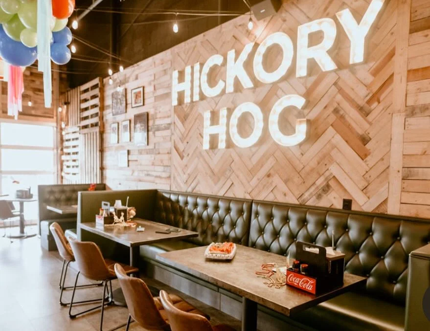 A cozy restaurant interior featuring a wooden wall with the name 'HICKORY HOG' illuminated. The seating area includes leather booths and modern tables.
