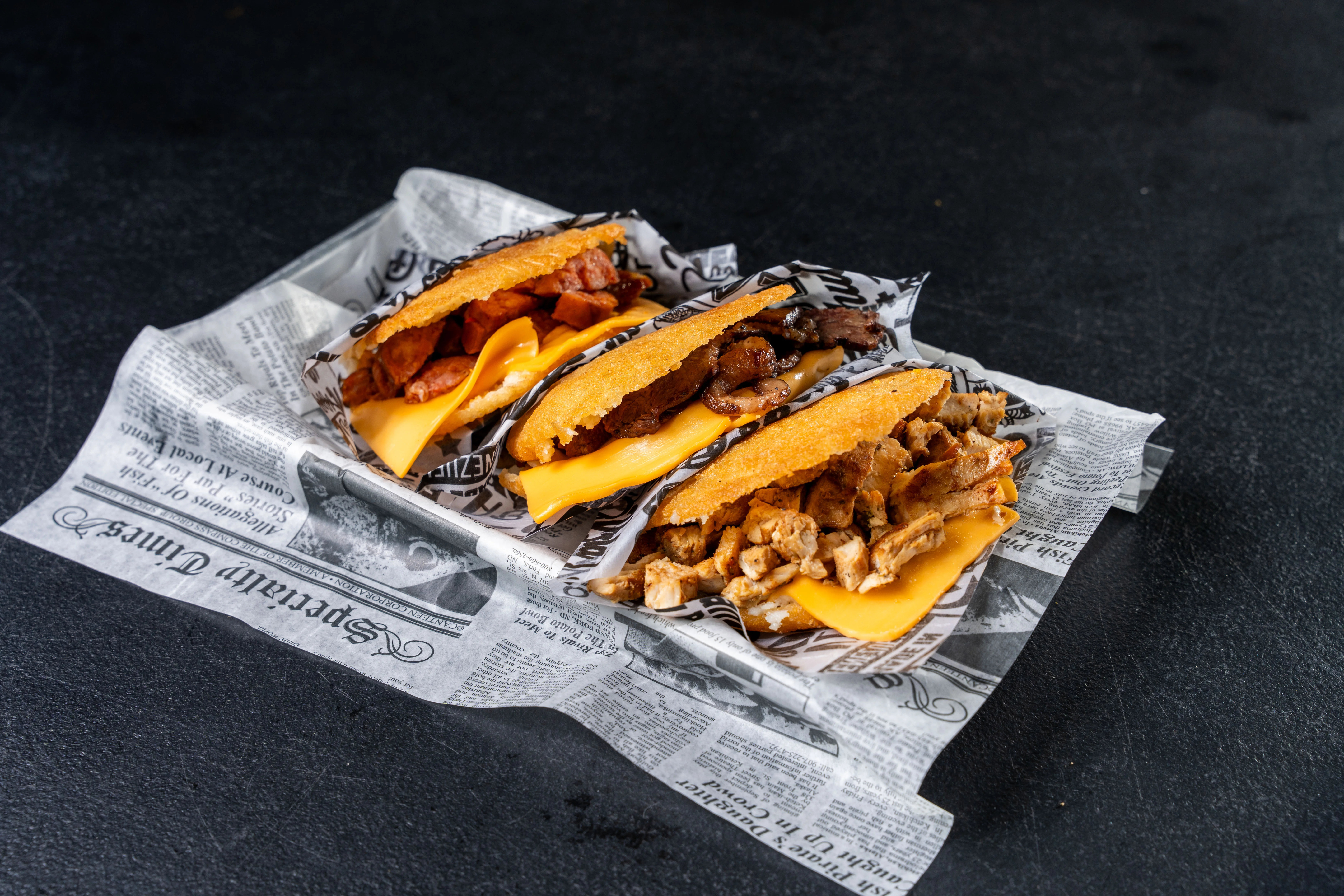 Three loaded tacos with cheese and meats on newspaper
