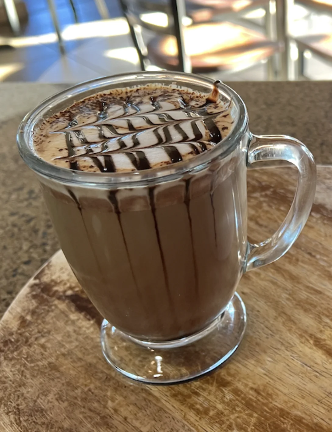 A hot chocolate beverage topped with a decorative swirl of chocolate syrup, served in a clear mug.