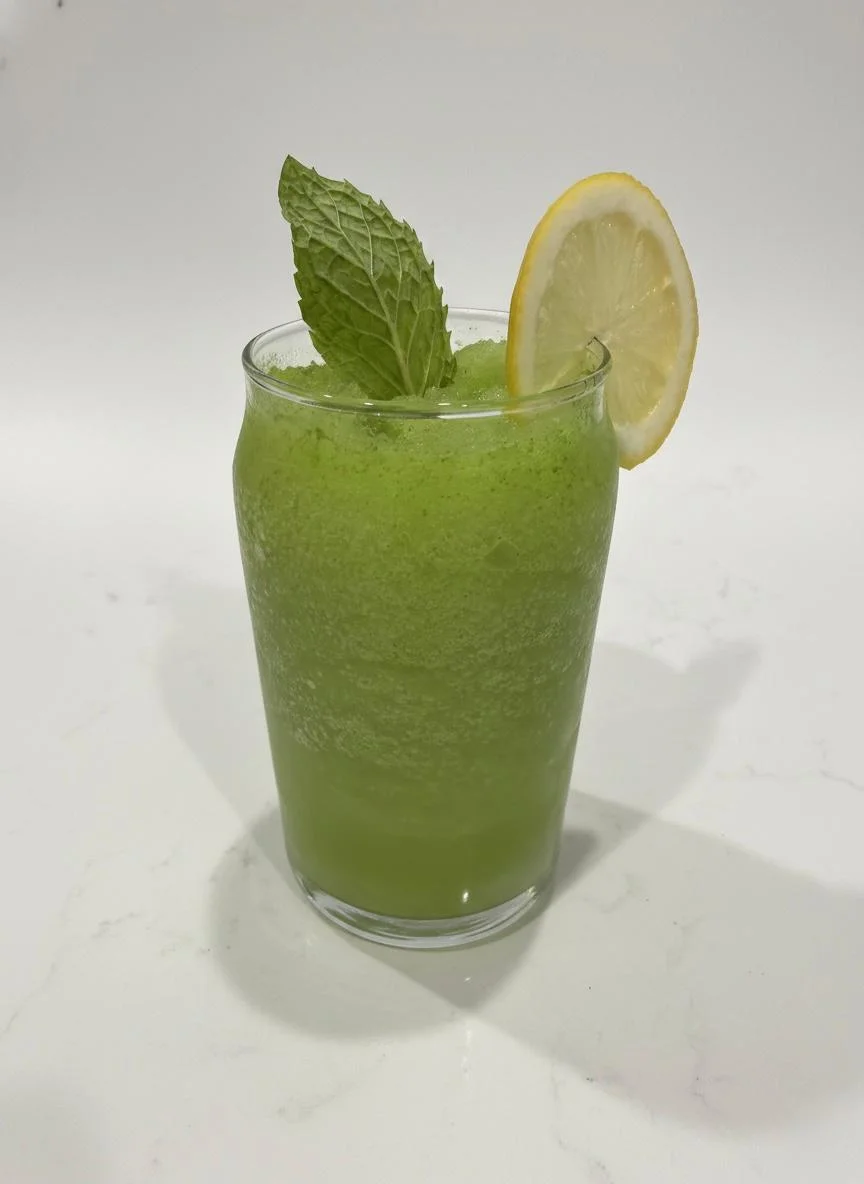A refreshing green drink served in a clear glass, garnished with a lemon slice and mint leaves.