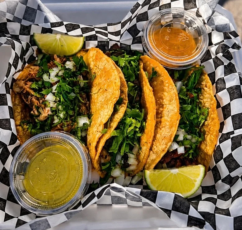 A box containing three tacos filled with meat and topped with onions and cilantro, served with lime and two containers of sauce.