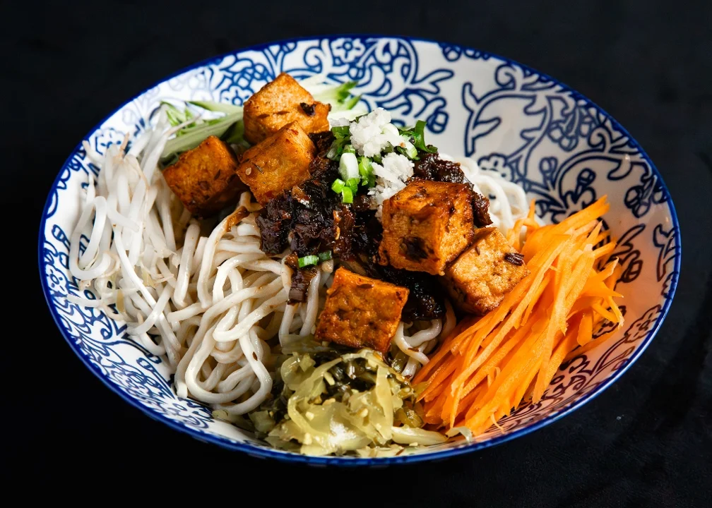 A bowl of noodles topped with fried tofu, scallions, minced garlic, a dark sauce, shredded carrots, and pickled greens.