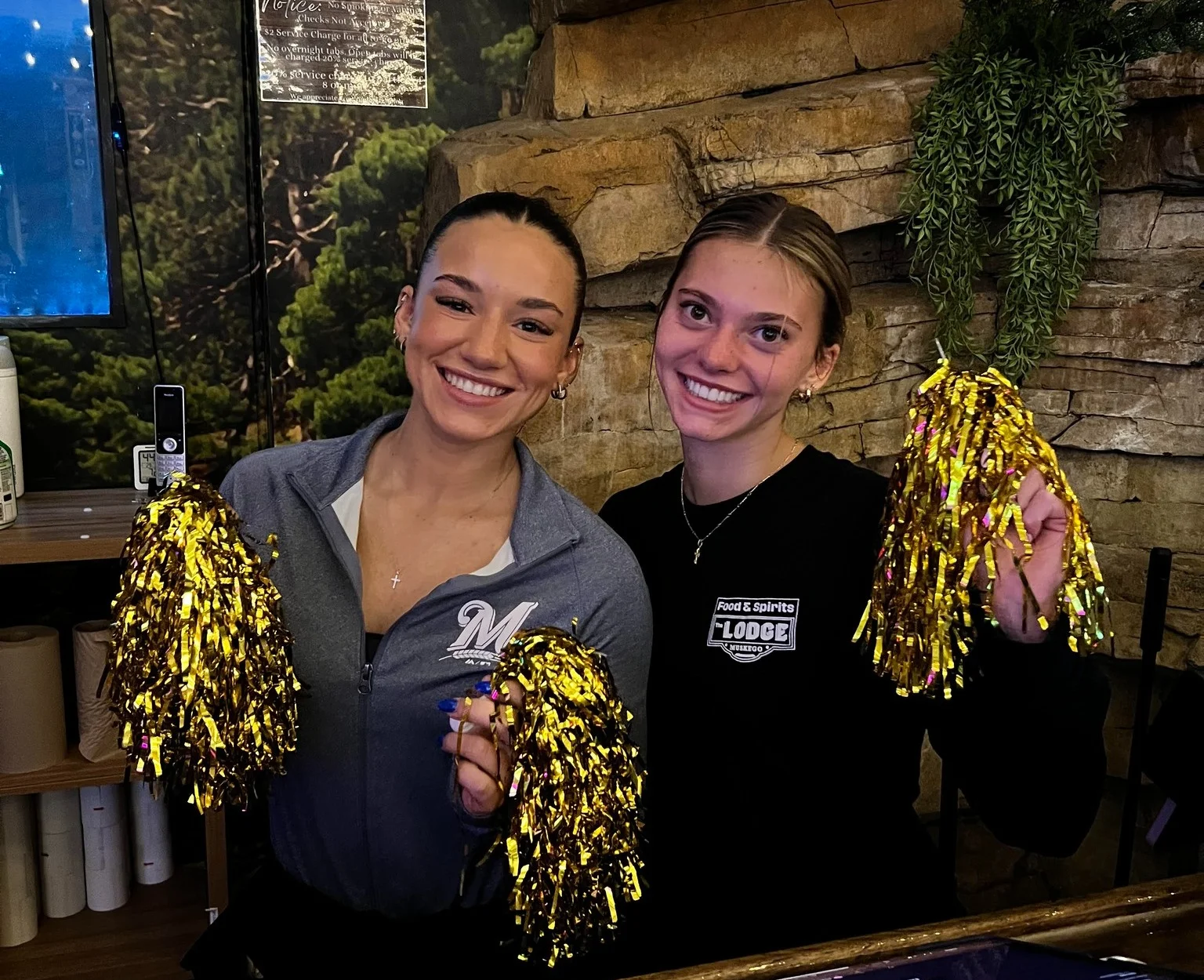 Two cheerful waitstaff holding gold pom-poms at a lodge bar