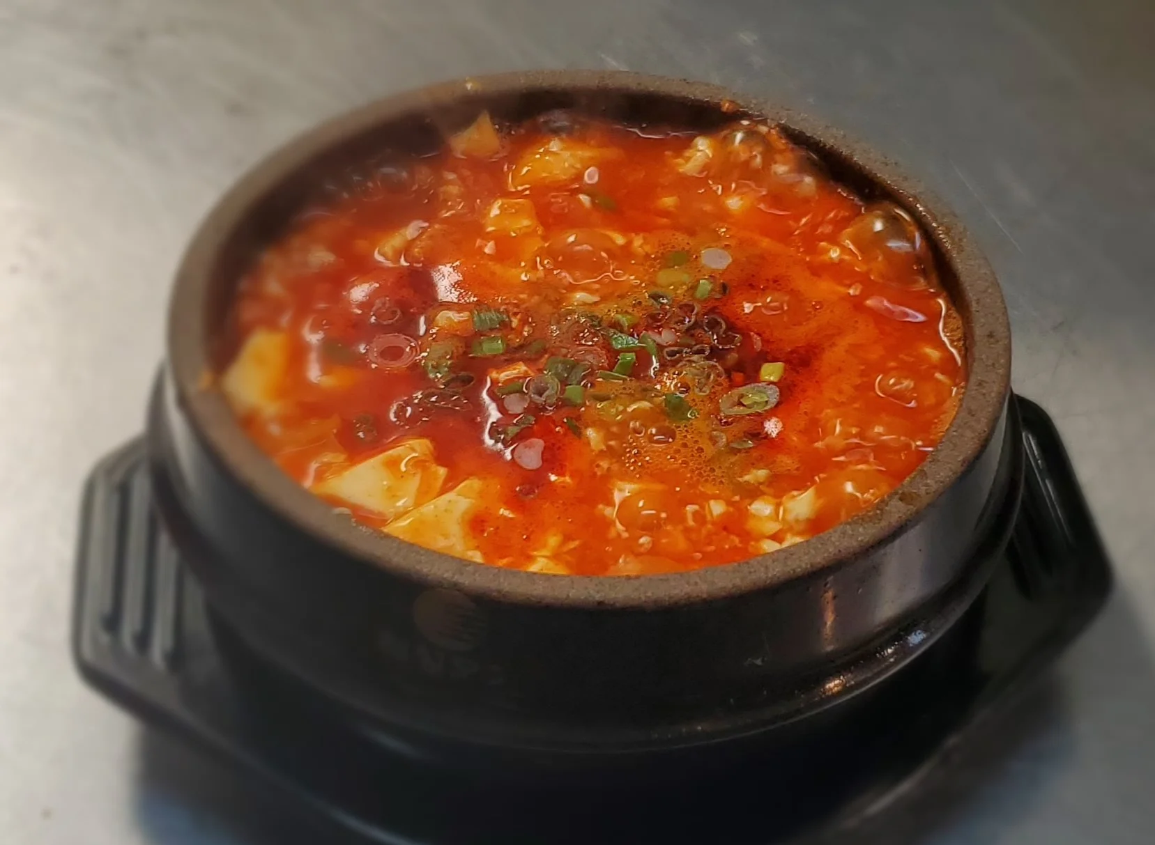 Korean sundubu jjigae (soft tofu stew) in black stone bowl with silky tofu, vegetables, and spicy red broth garnished with scallions