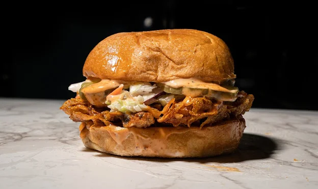 A hearty burger filled with shredded meat, fries, and cheese, all topped with a creamy sauce, placed on a brown bun.