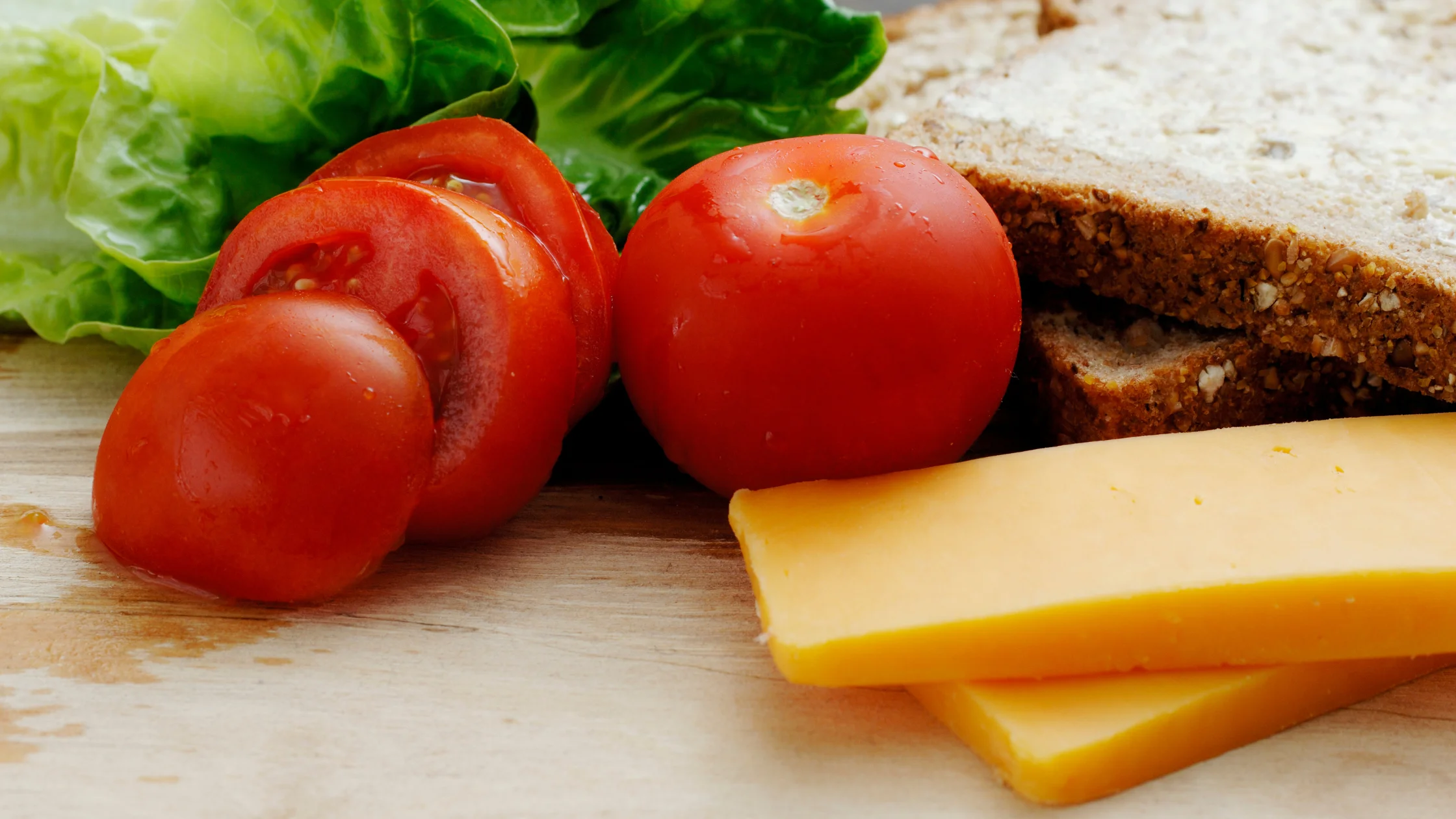 Fresh sandwich ingredients including sliced tomatoes, whole tomatoes, leafy romaine lettuce, cheddar cheese slices, and whole grain bread on a wooden surface
