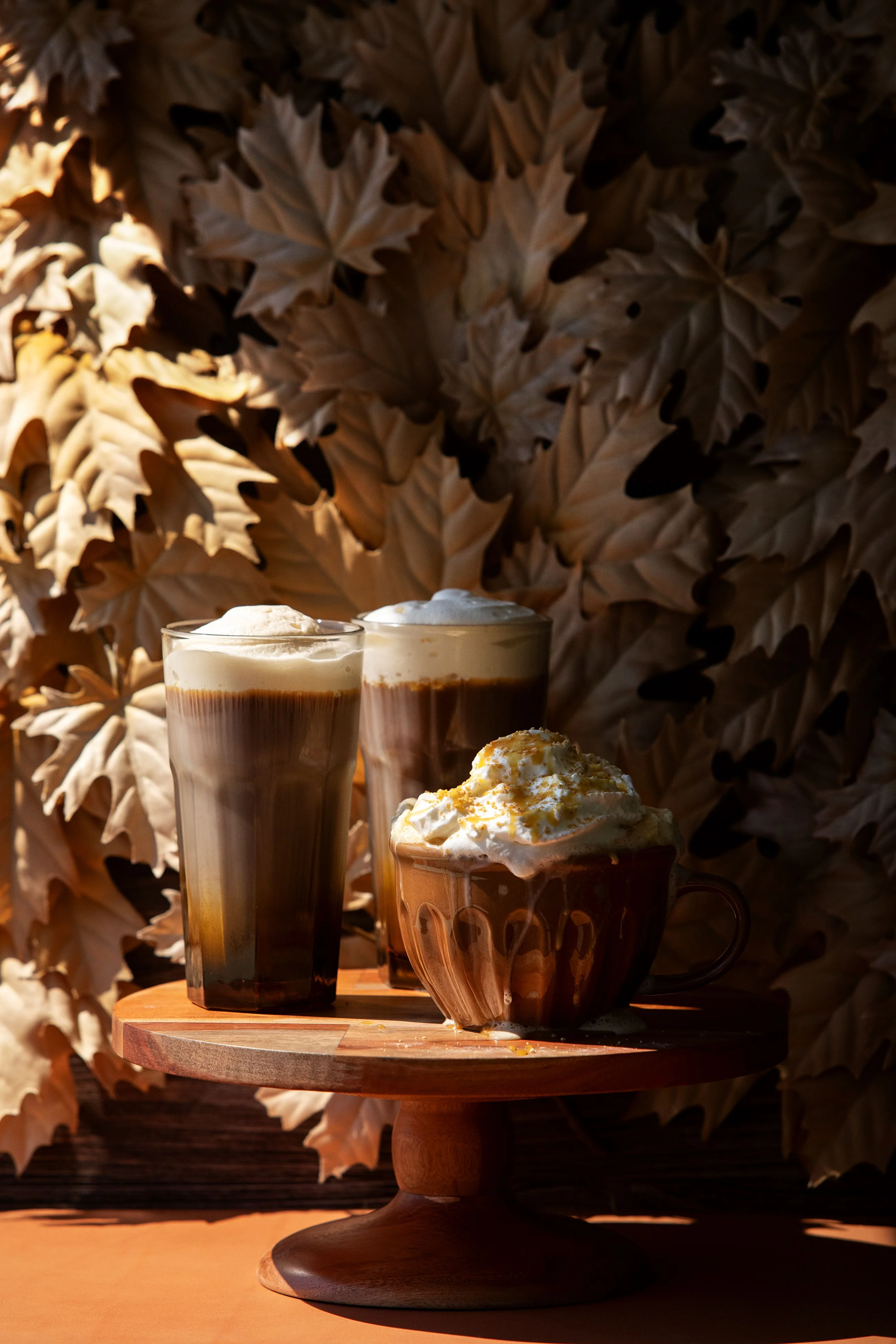 Three frothy coffee drinks rest on a wooden stand against a backdrop of autumn leaves. The scene conveys a warm, cozy fall atmosphere.