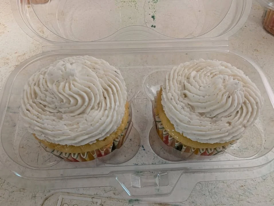 Two vanilla cupcakes with generous swirls of white icing, displayed in a transparent plastic container.