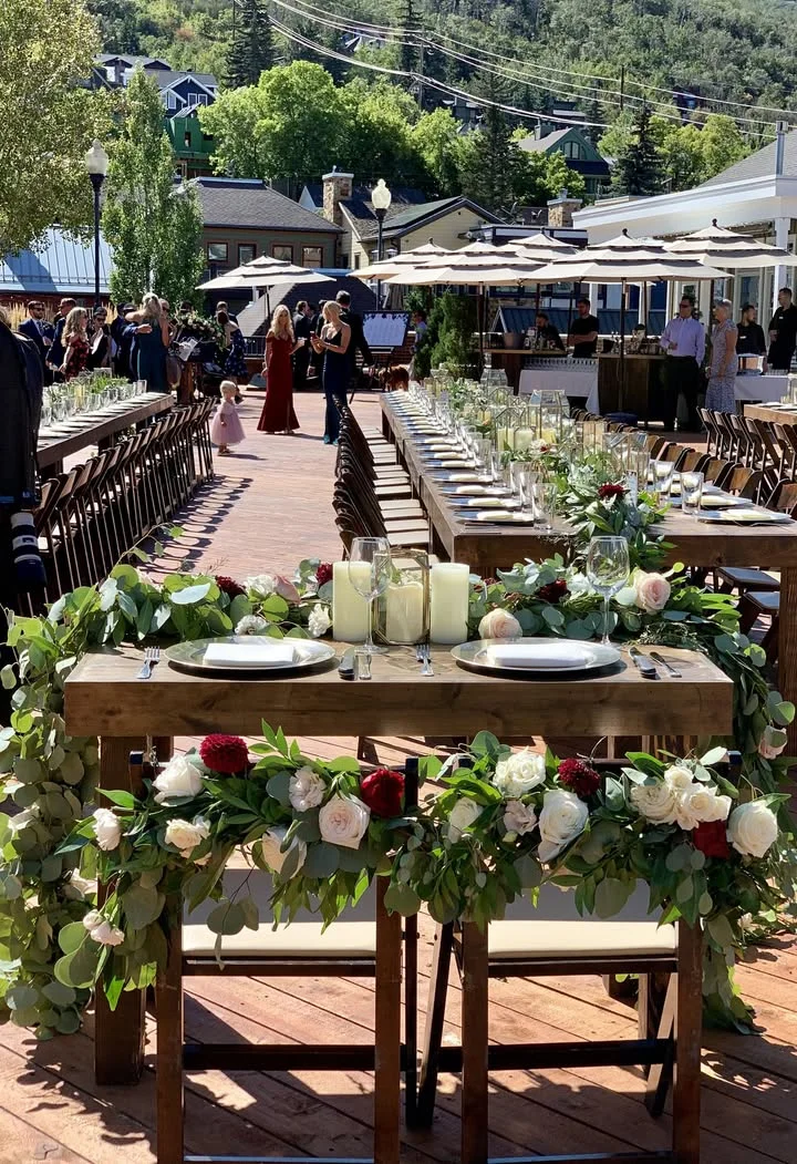 An elegant outdoor dining setup with long tables adorned with floral arrangements and glassware, set for an event.