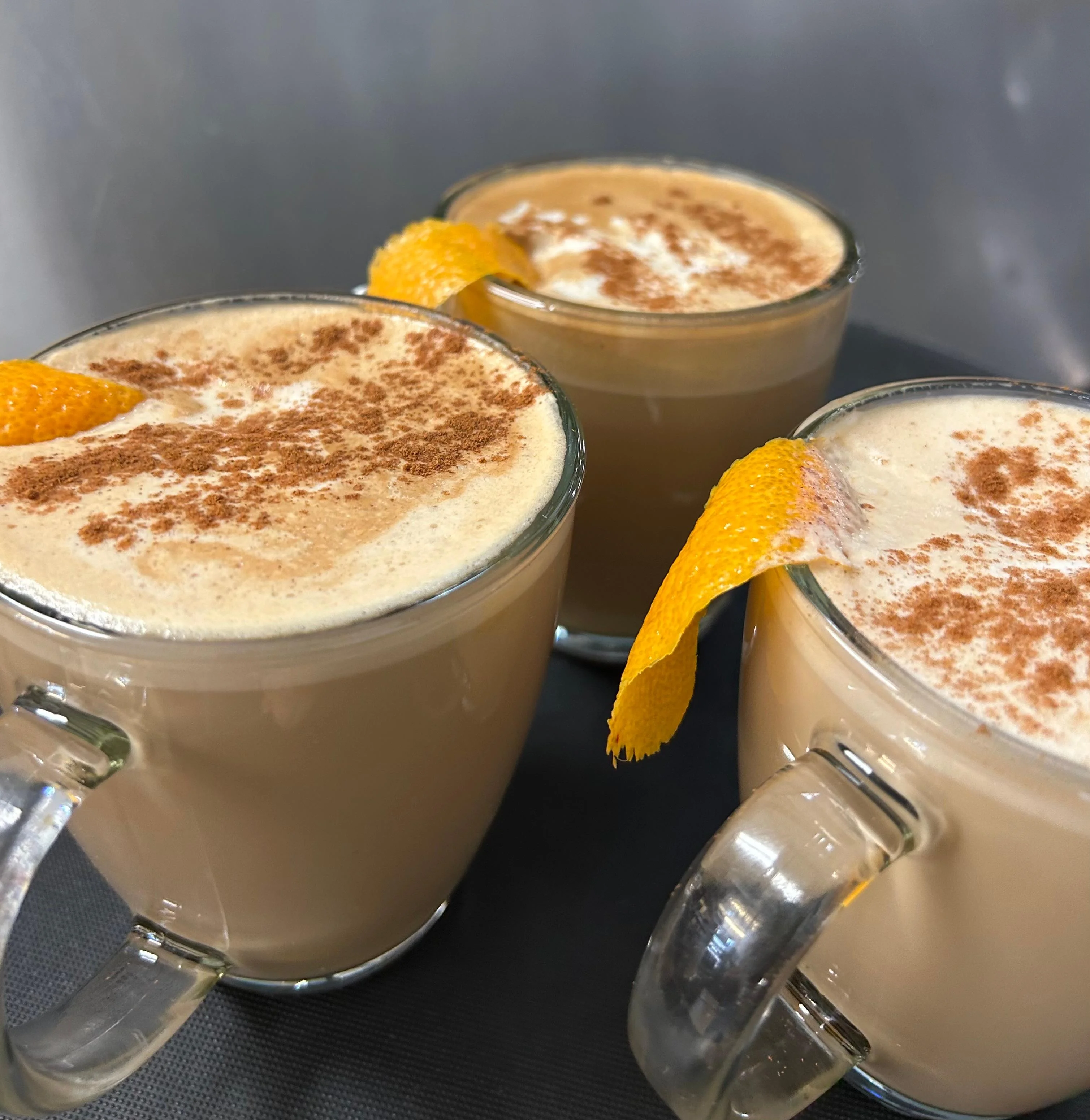 Three cups of creamy beverages topped with cinnamon and garnished with orange peels, served on a dark surface.