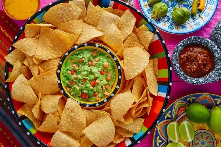 A vibrant display of tortilla chips served in a colorful bowl, accompanied by a green guacamole dip topped with diced tomatoes, alongside lime wedges and salsa.