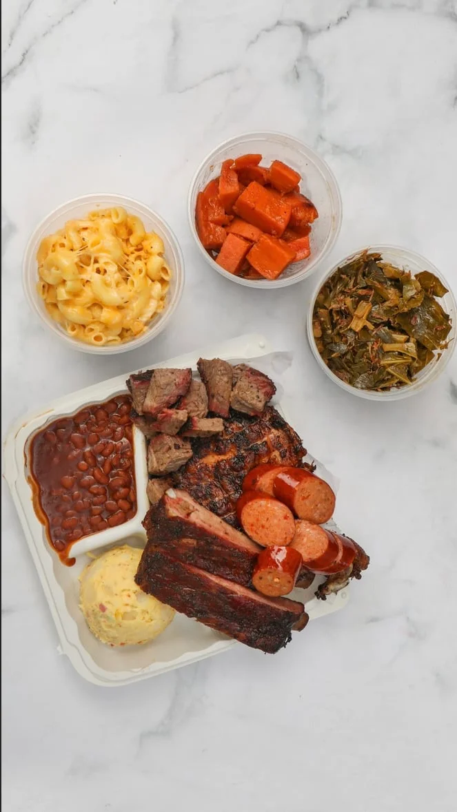An assorted BBQ platter with various meats, baked beans, potato salad, and additional sides in separate containers.