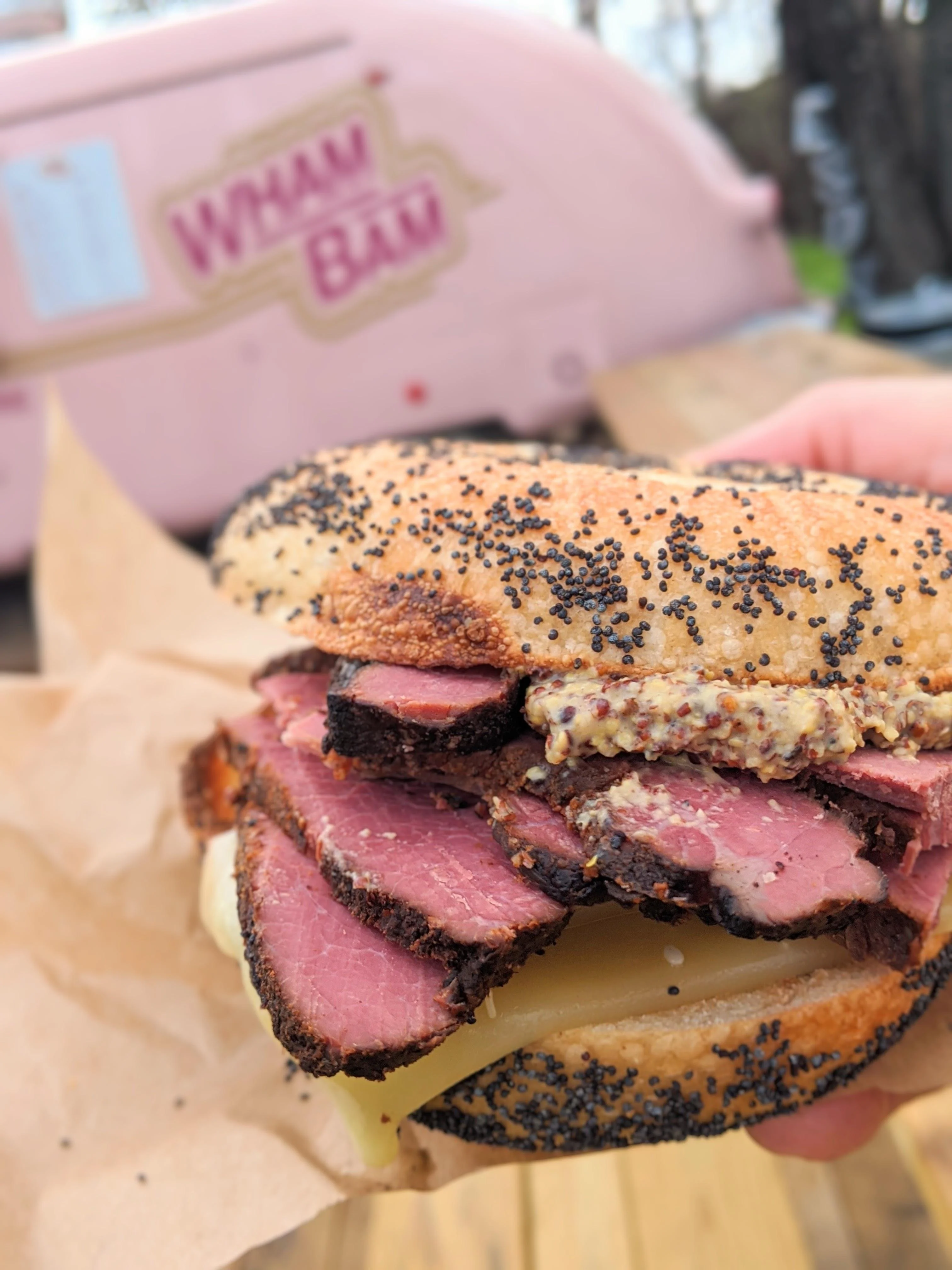 A poppy seed bagel sandwich filled with layers of smoked meat and mustard. The sandwich is held in hand with a pink food truck in the background.