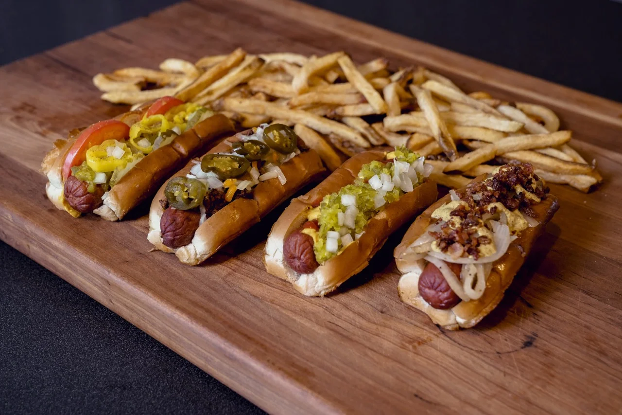 A wooden board featuring four gourmet hot dogs topped with a variety of ingredients and a side of golden fries.