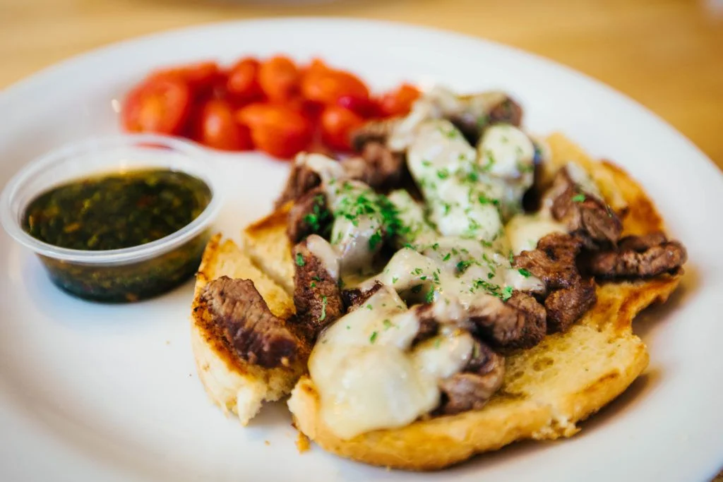 A savory dish featuring grilled steak served on toasted bread with melted cheese and a side of fresh tomatoes and green sauce.