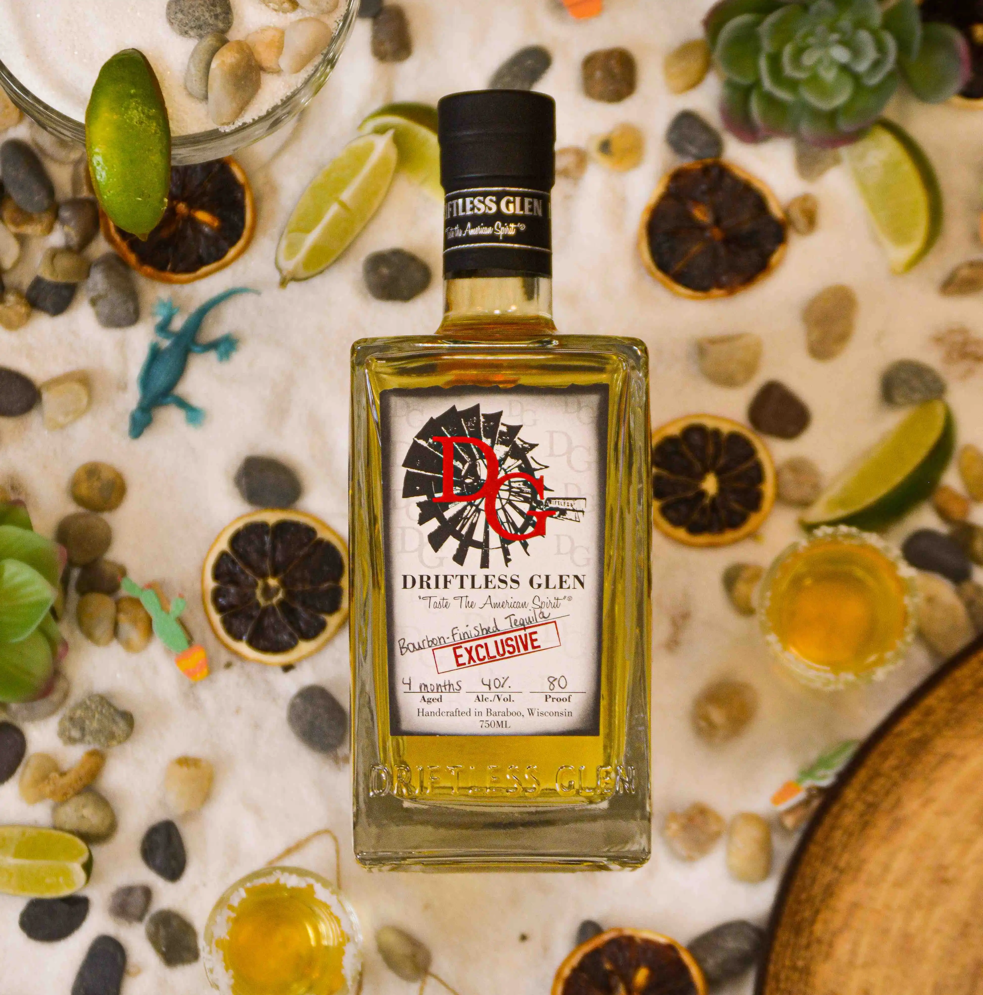 A bottle of Bourbon-Finished Tequila sits on a sandy backdrop with limes, cactuses, and rocks. 