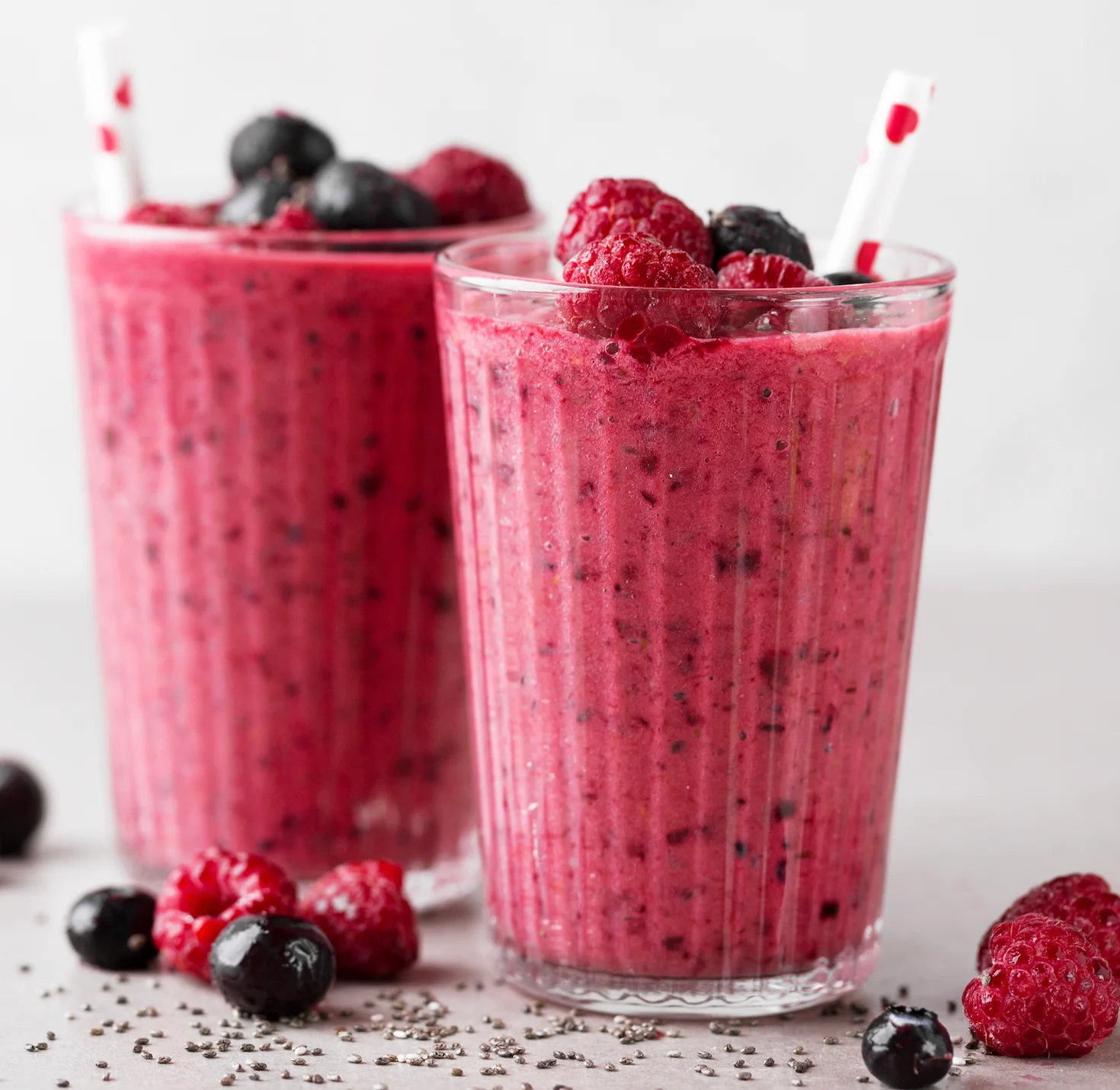 Two tall glasses of berry smoothie, decorated with raspberries and blueberries, accompanied by chia seeds on the table.