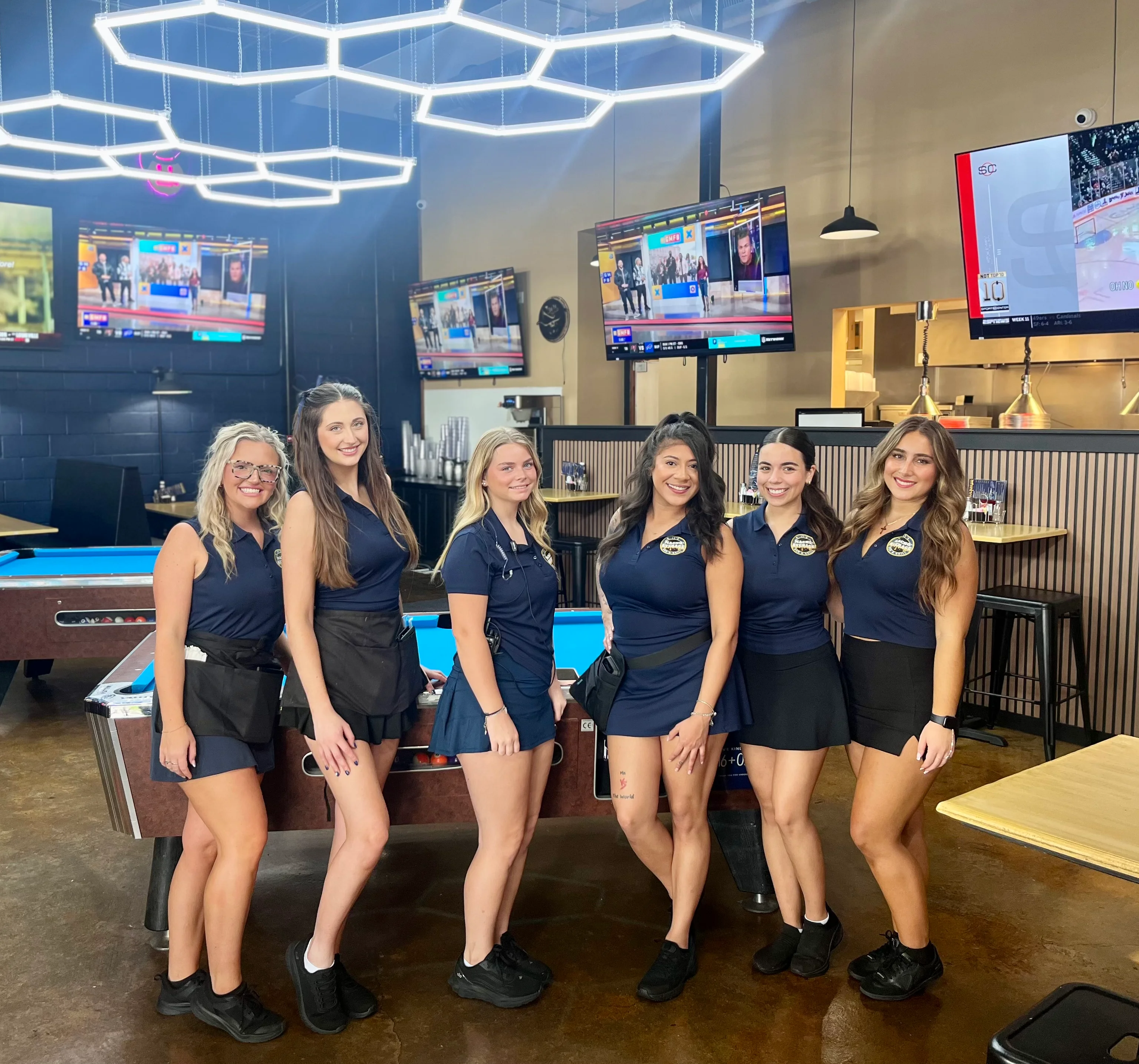 A lively indoor entertainment area featuring pool tables, people socializing, and multiple screens showing sports broadcasts. The atmosphere is vibrant with modern lighting elements.
