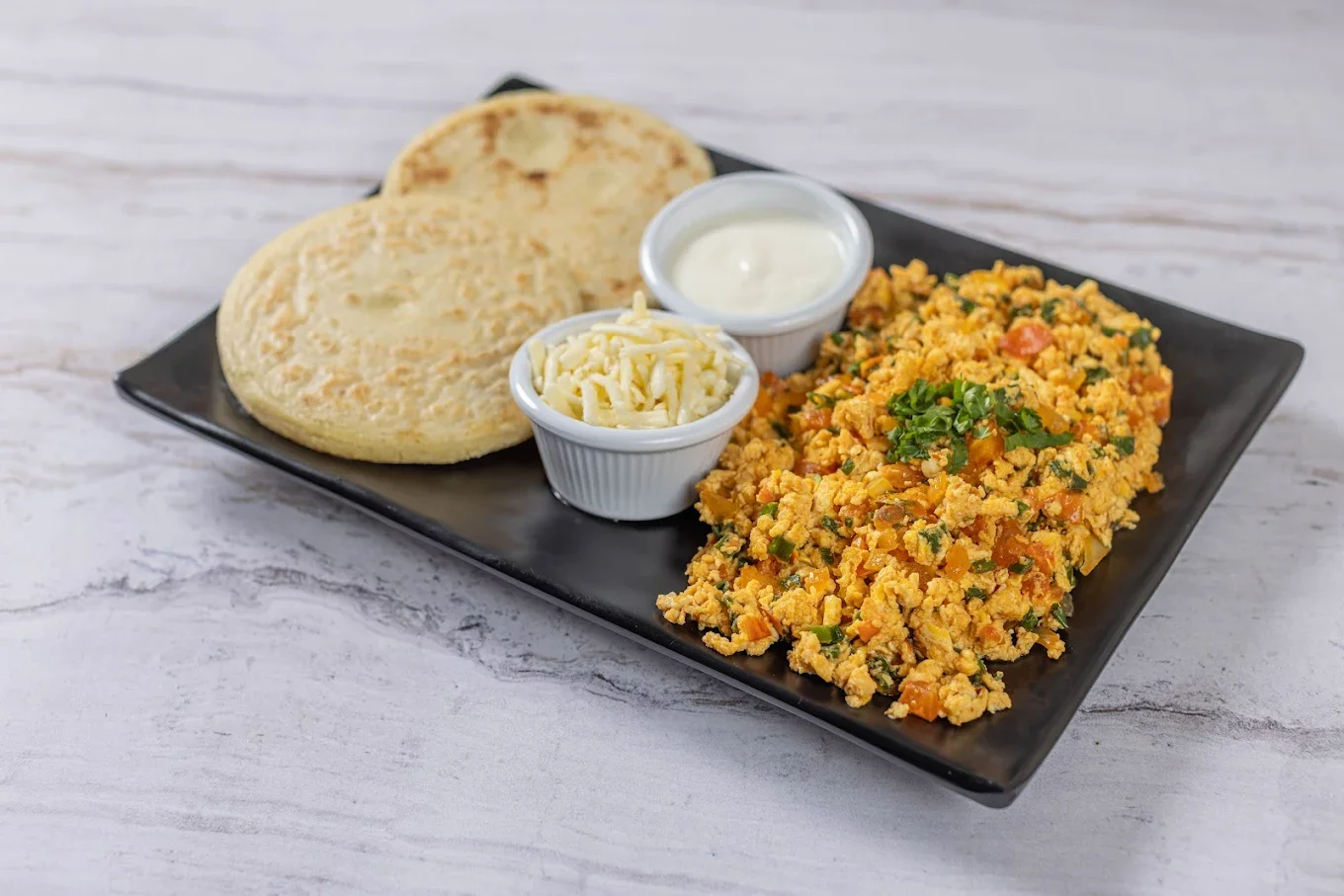 A traditional Venezuelan breakfast plate at El Budare Cafe, featuring perico (scrambled eggs), fresh arepas, shredded white cheese, and nata (cream).