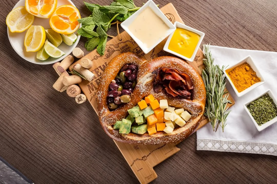 A large pretzel arranged with various toppings including olives, bacon, and cheese, alongside an assortment of fresh fruits and sauces on a wooden board.