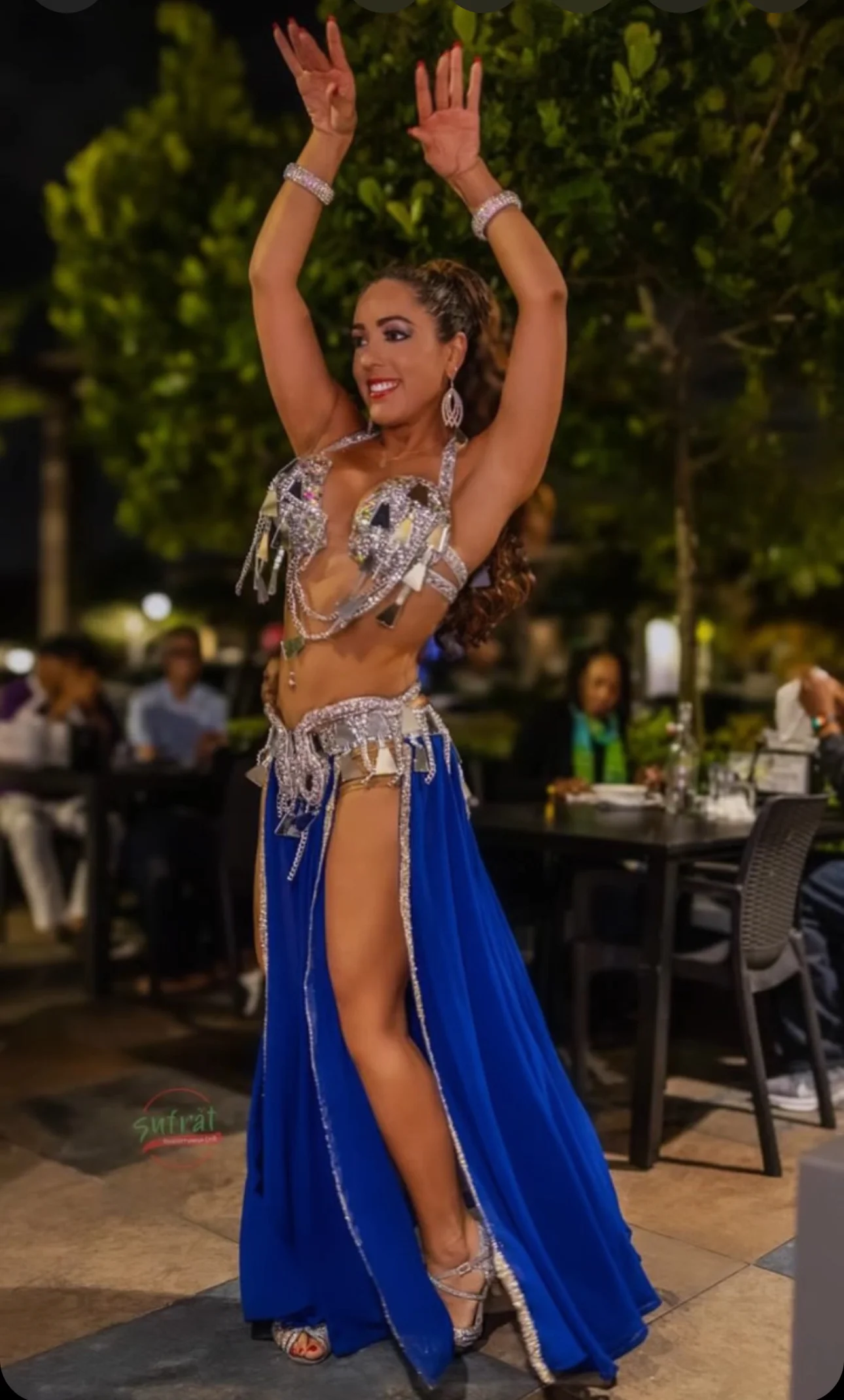 Dancer in an ornate silver and blue costume performs joyfully at an outdoor restaurant at night, with blurred diners in the background.