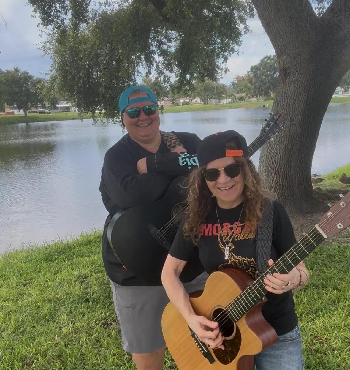 Two musicians by a lake, one playing guitar and the other smiling.