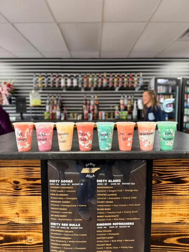 An assortment of colorful beverages in clear cups on a countertop, with a menu board in the foreground. The drinks are creatively named and arranged neatly.