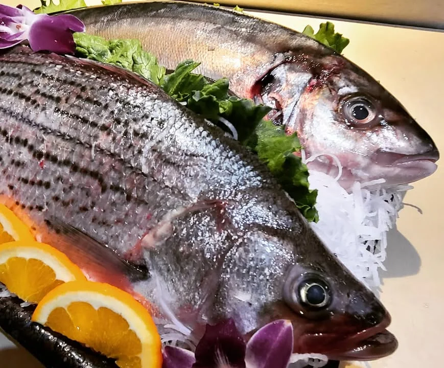 Two whole fish displayed on a bed of lettuce, garnished with orange slices and flowers.