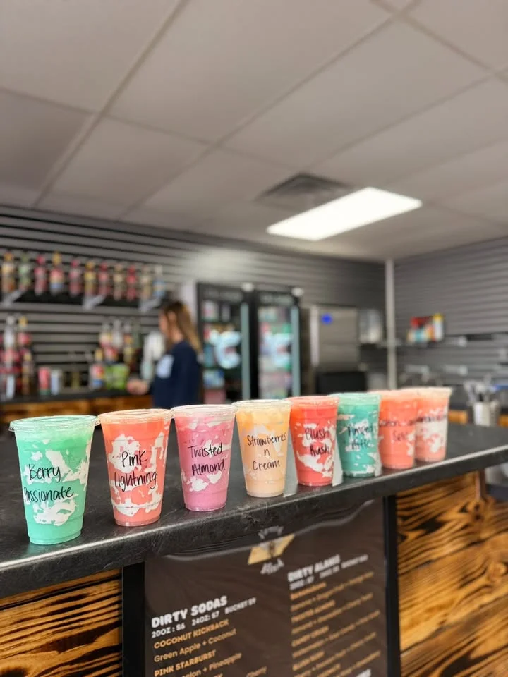 A variety of colorful drinks displayed in clear cups on a counter, each labeled with different names. The background shows a well-organized shop with various beverage options.