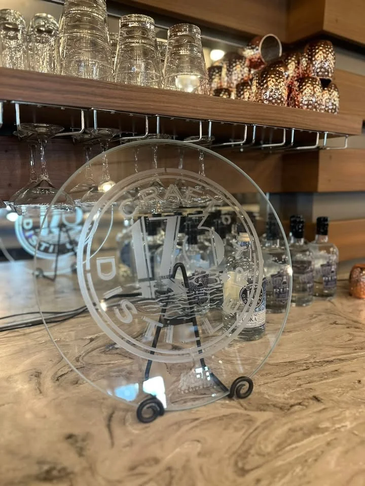 An elegant shelf displaying glassware and a decorative acrylic plate with a logo inside a modern bar or distillery.