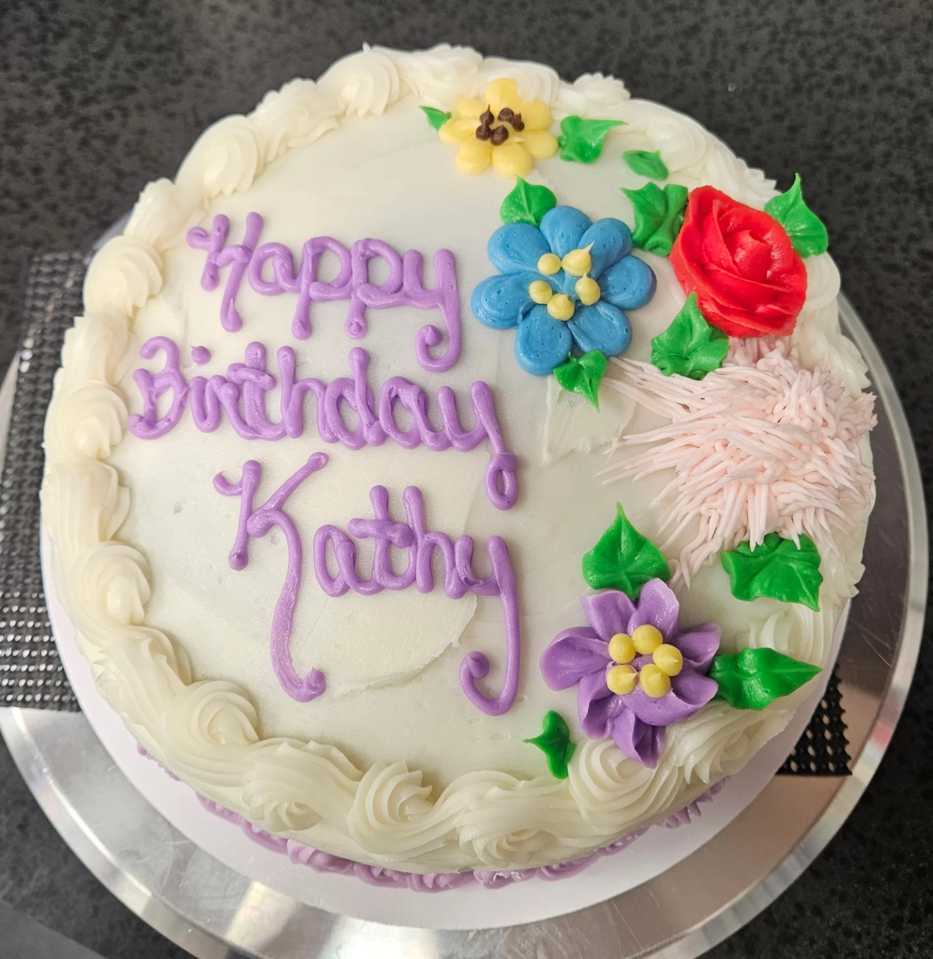 Round cake with colorful flowers on the right side & on the left says "Happy Birthday Kathy"