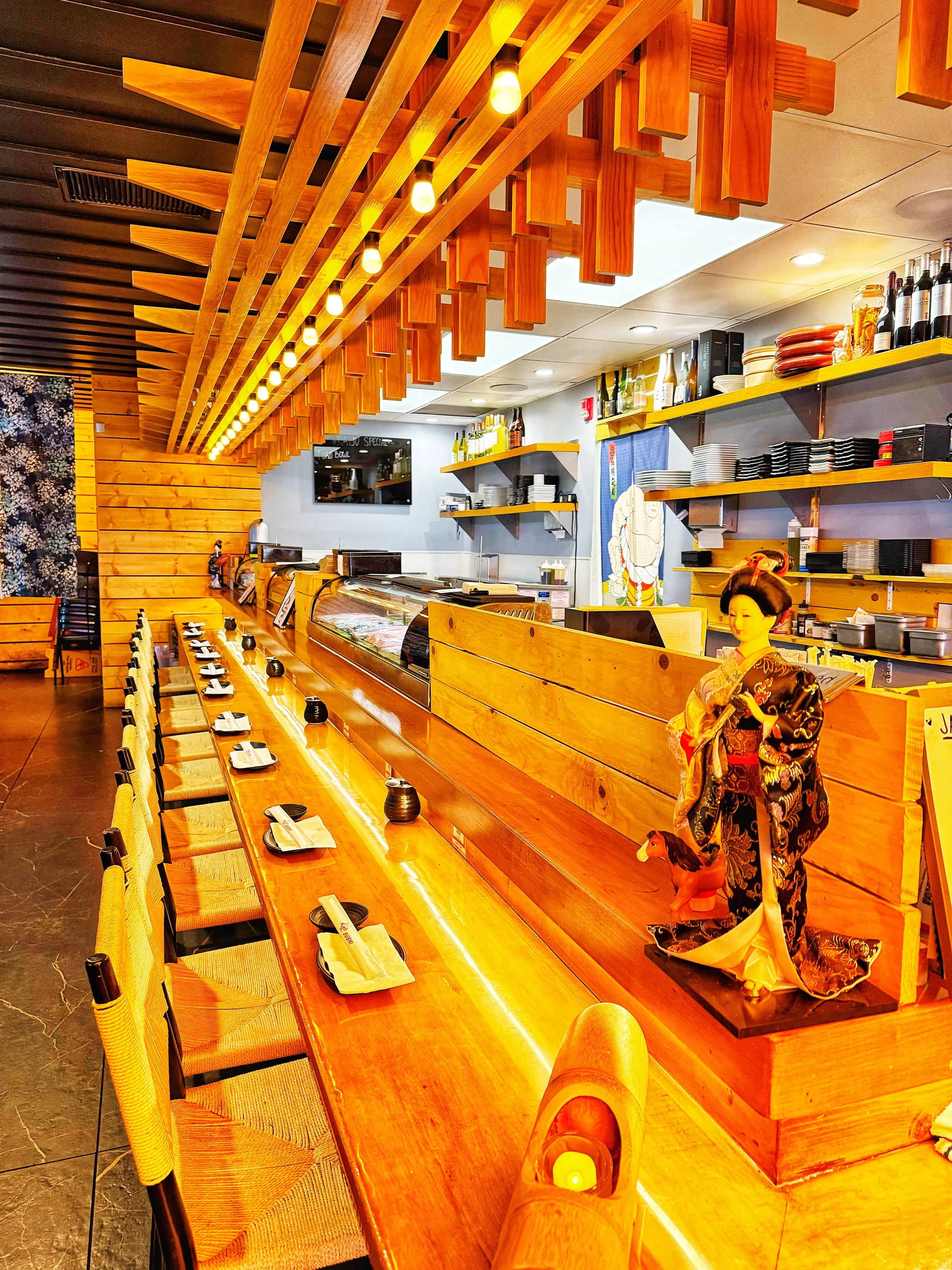 A modern sushi restaurant interior featuring a long wooden counter with seat arrangements. Decorative lights and traditional elements are visible in the ambiance.