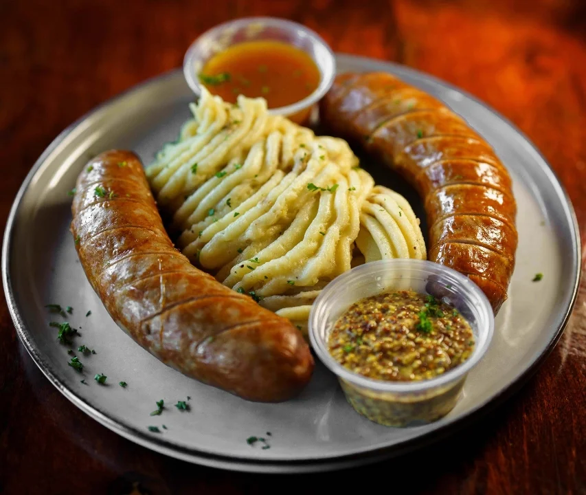 A plate featuring two sausage links paired with a serving of creamy mashed potatoes, accompanied by mustard and gravy in small cups.
