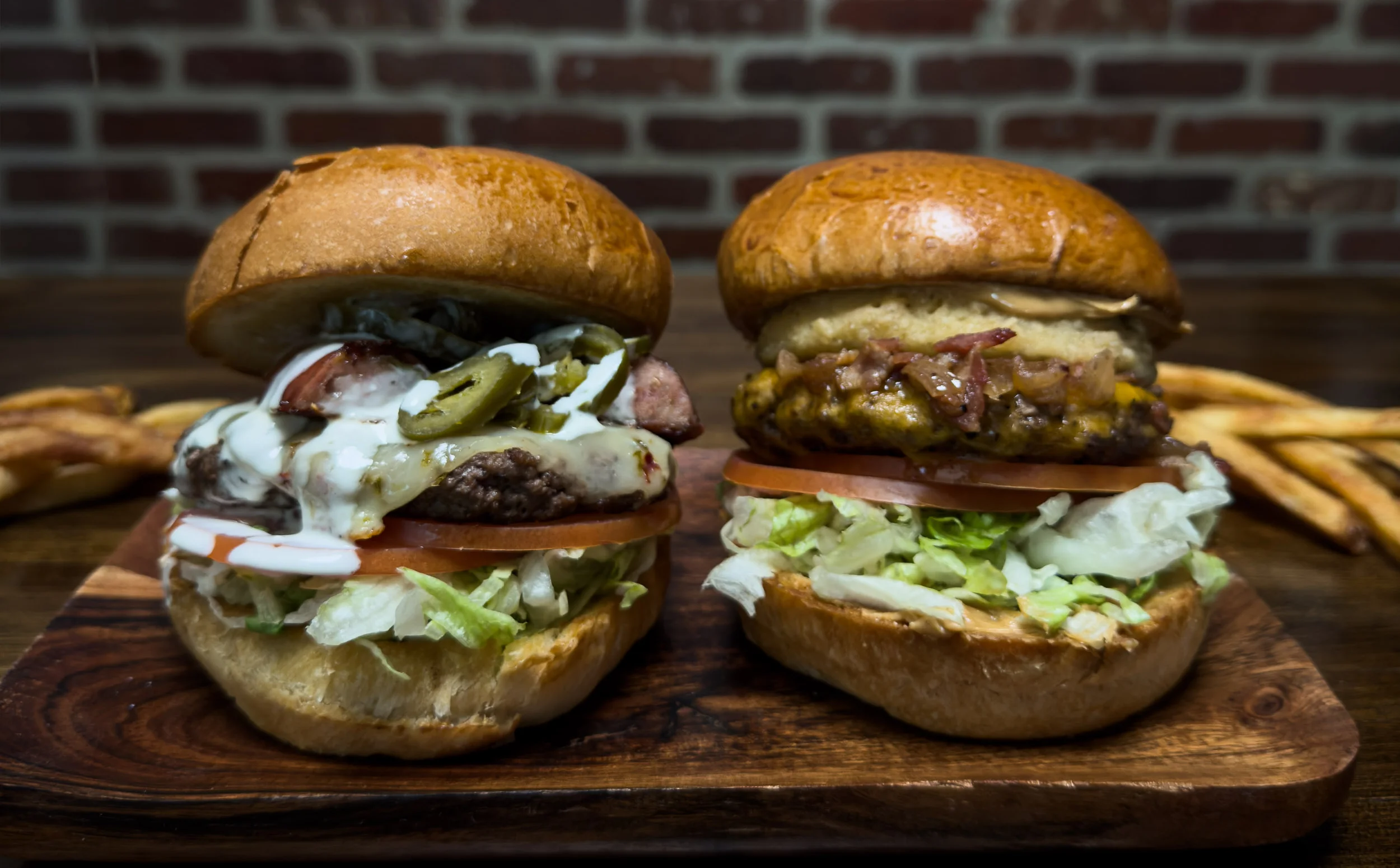 Two gourmet burgers with toppings on a wooden board, served with fries.