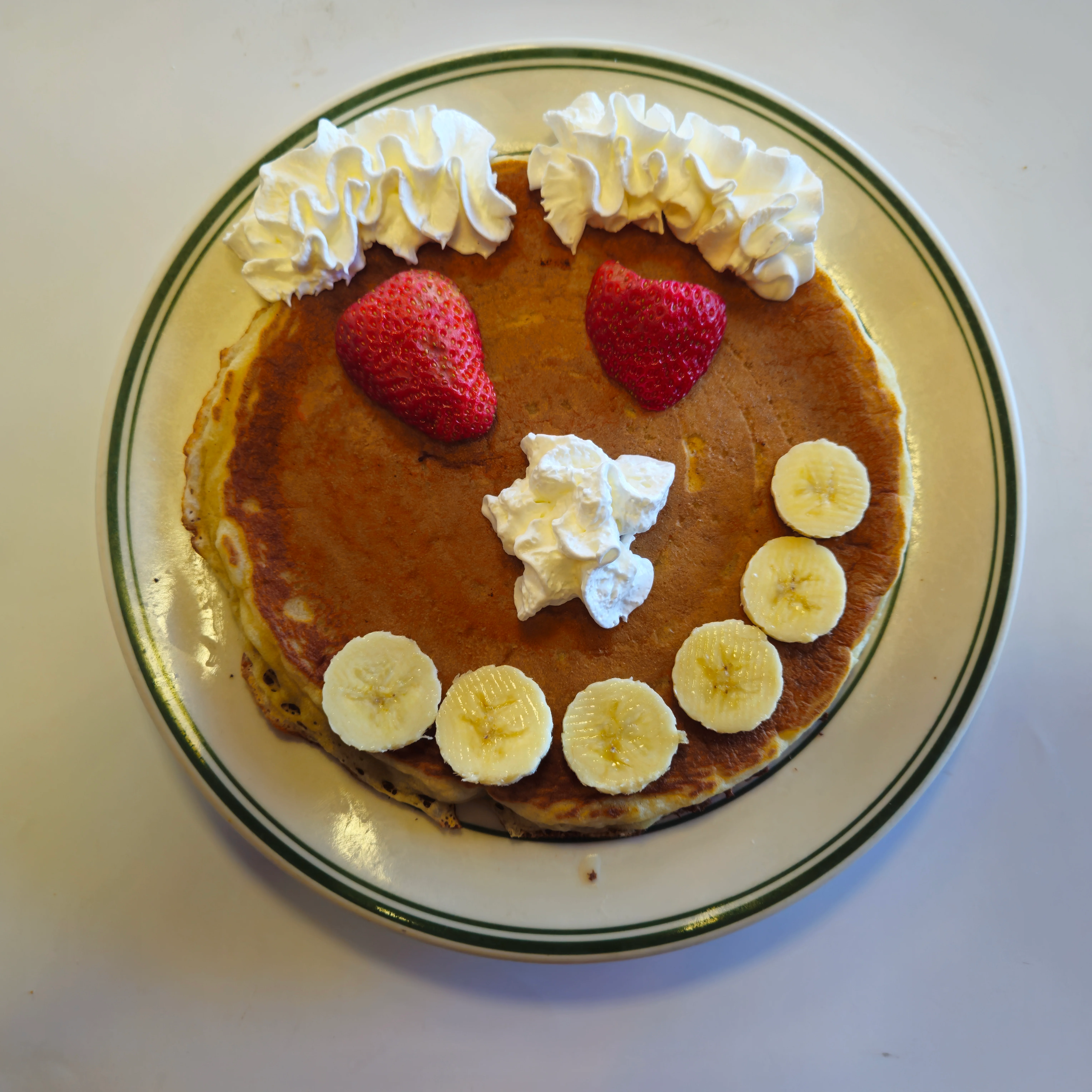 A creative pancake presentation featuring whipped cream, strawberries, and banana slices arranged to form a smiley face.