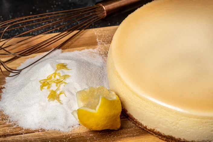 Lemon cheesecake with sugar, lemon zest, and a whisk on a wooden board.