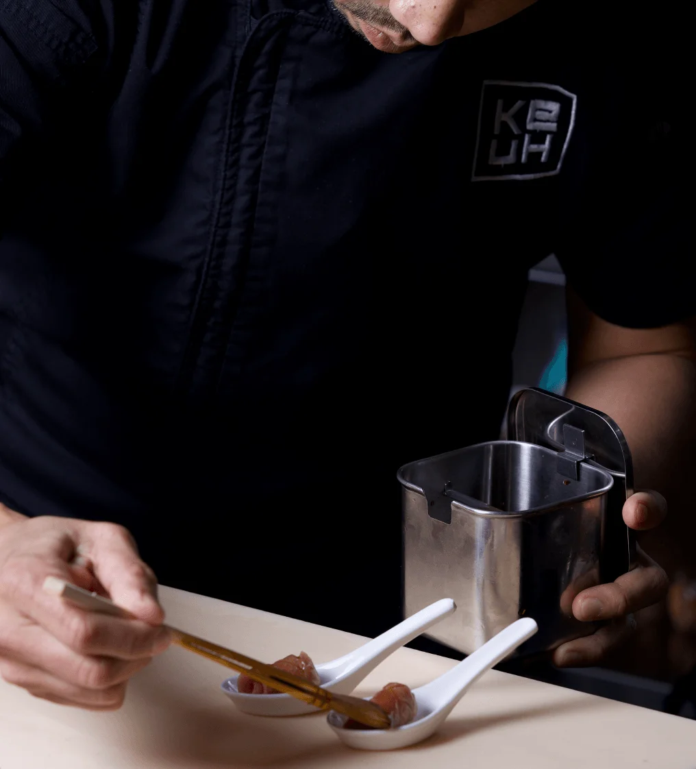 A chef wearing black gloves carefully garnishing a plate of fresh yellowtail nigiri with microgreens and jalapeño.